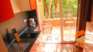 Une cuisine moderne équipée d’un évier, d’une machine à café et de chaises en bois, située près d’une porte-fenêtre coulissante donnant sur un balcon avec vue sur les arbres.
