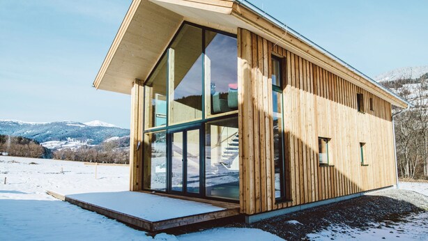 Een moderne houten blokhut met grote glasramen, een houten terras en uitzicht op besneeuwde bergen.