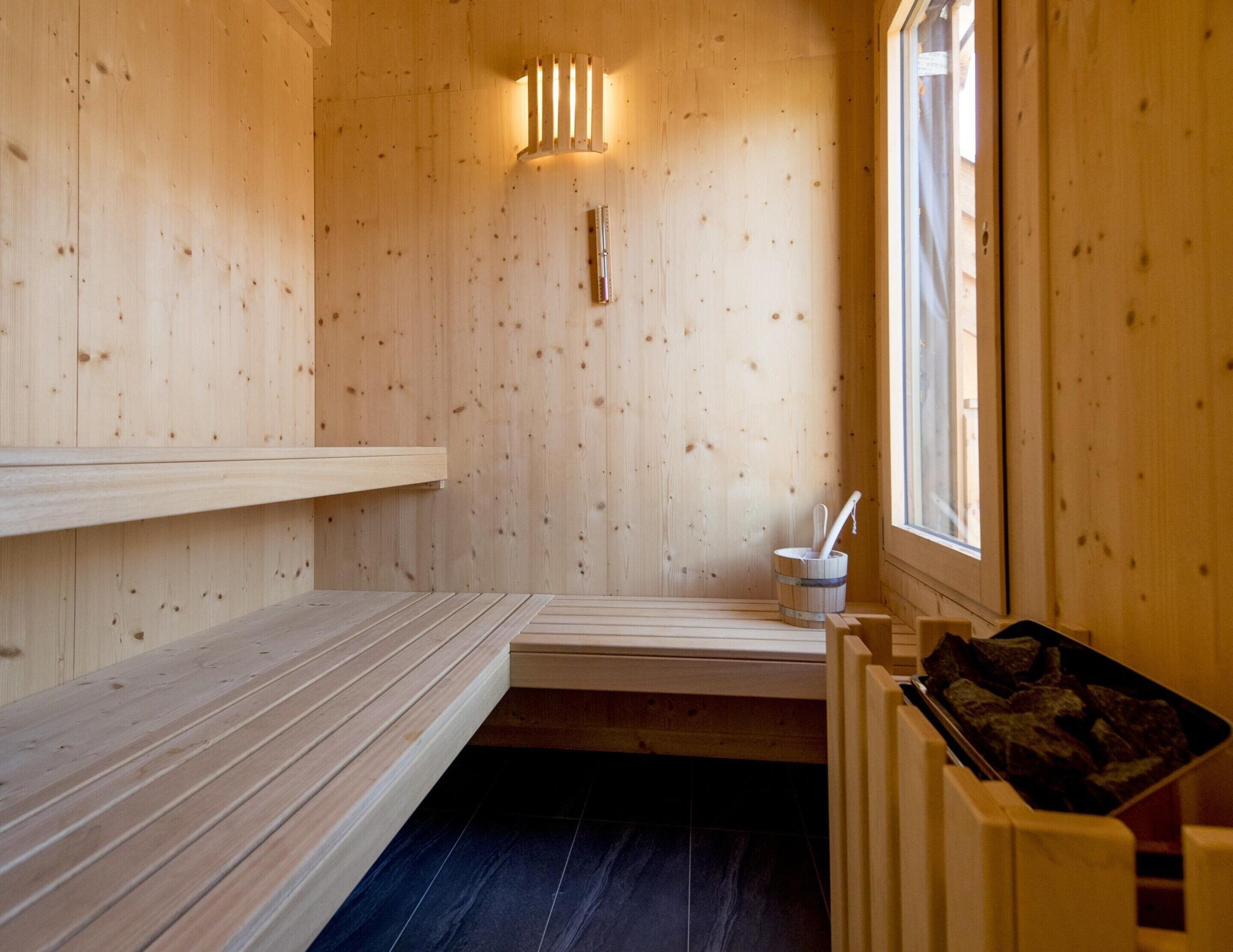 A wooden sauna room with benches, a wall-mounted light, a window, and a bucket with a brush.