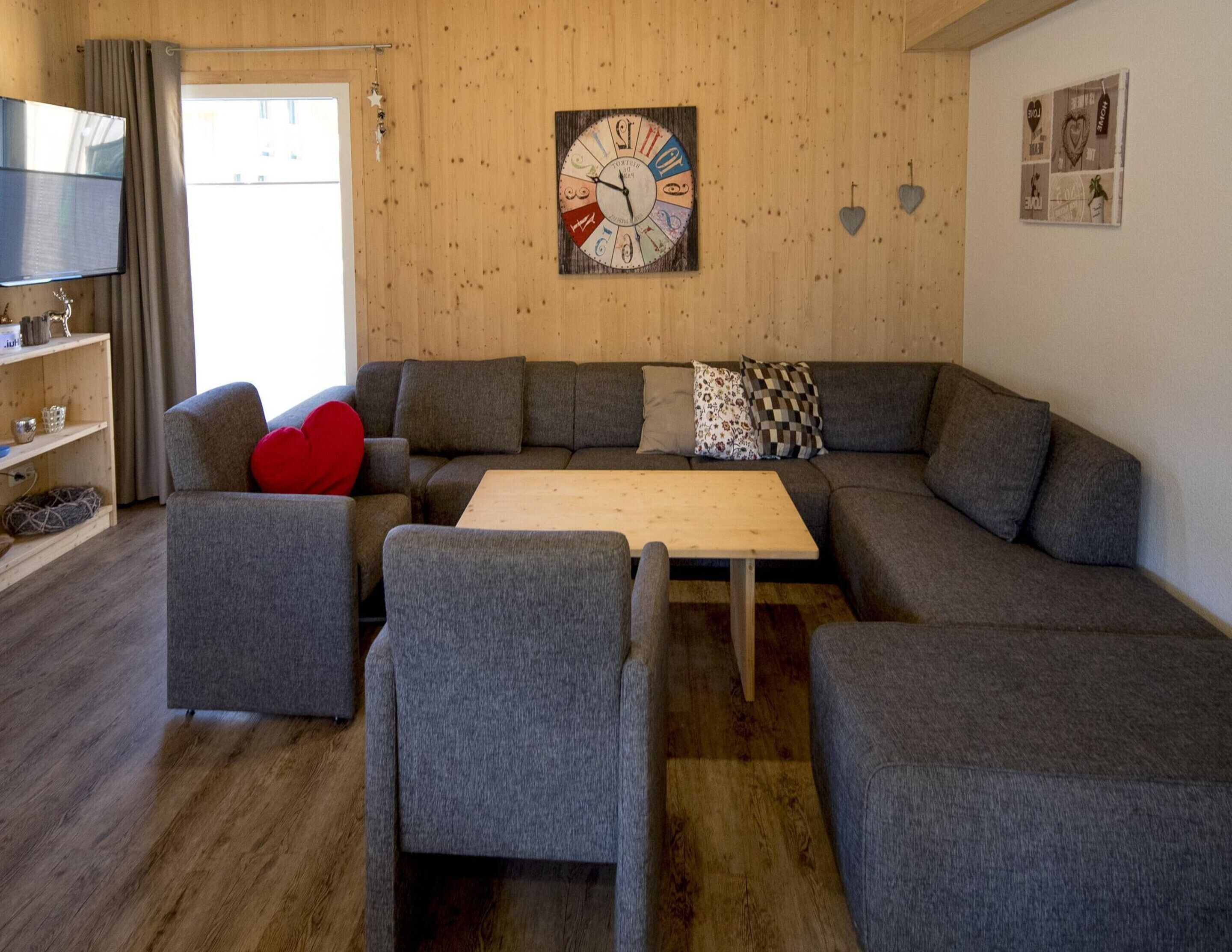 Un salon moderne comprenant un canapé d’angle, deux fauteuils, une table basse en bois et une horloge murale.