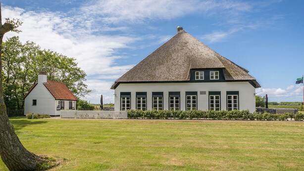 Ett vitt hus med ett halmtak och ett mindre vitt hus med ett rött tak i bakgrunden.