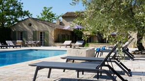 Un espace piscine avec des chaises longues et des parasols, un bâtiment en pierre et des arbres.