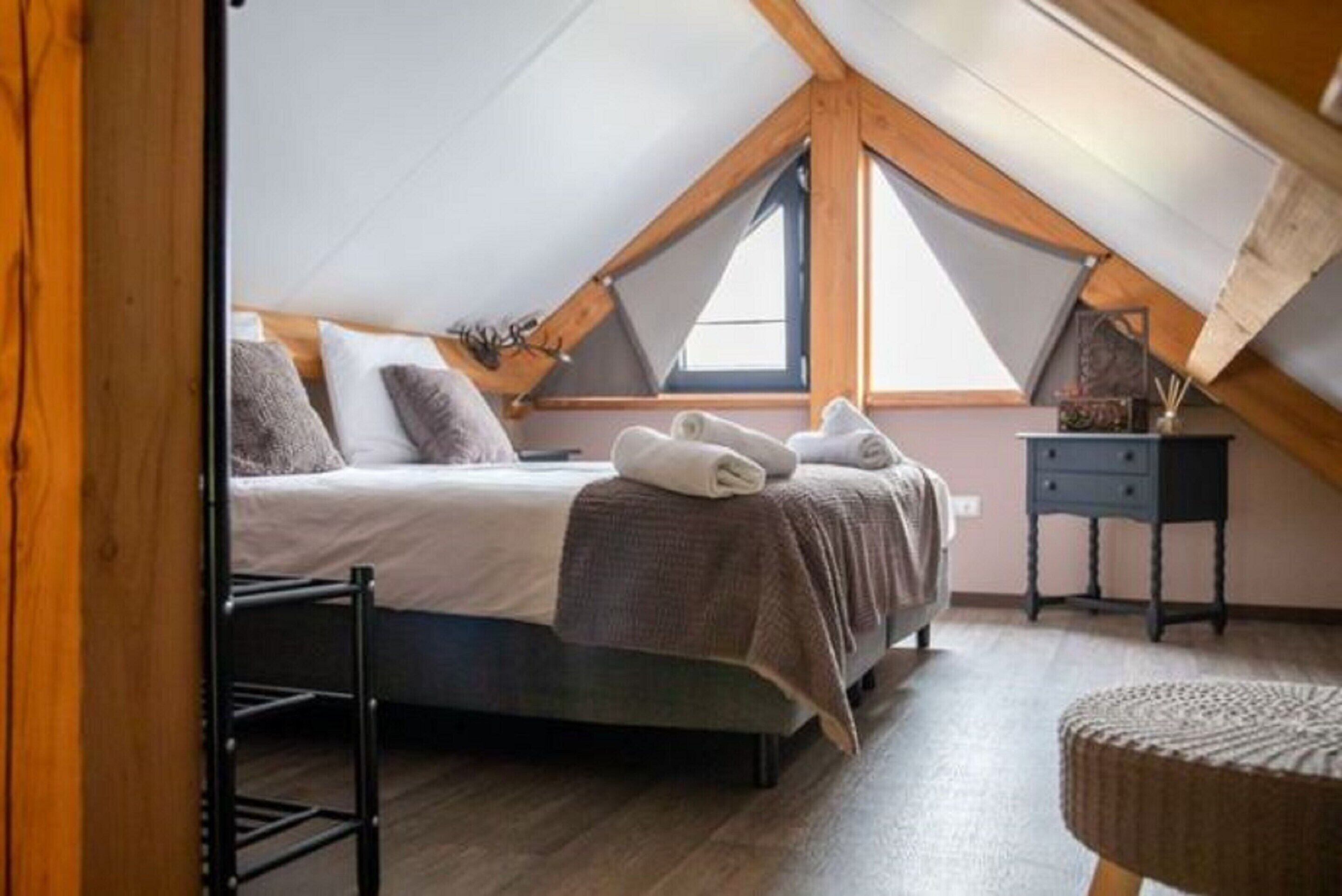 A bedroom with a loft-style ceiling, a bed with pillows and a blanket, a small bedside table, and a chair.