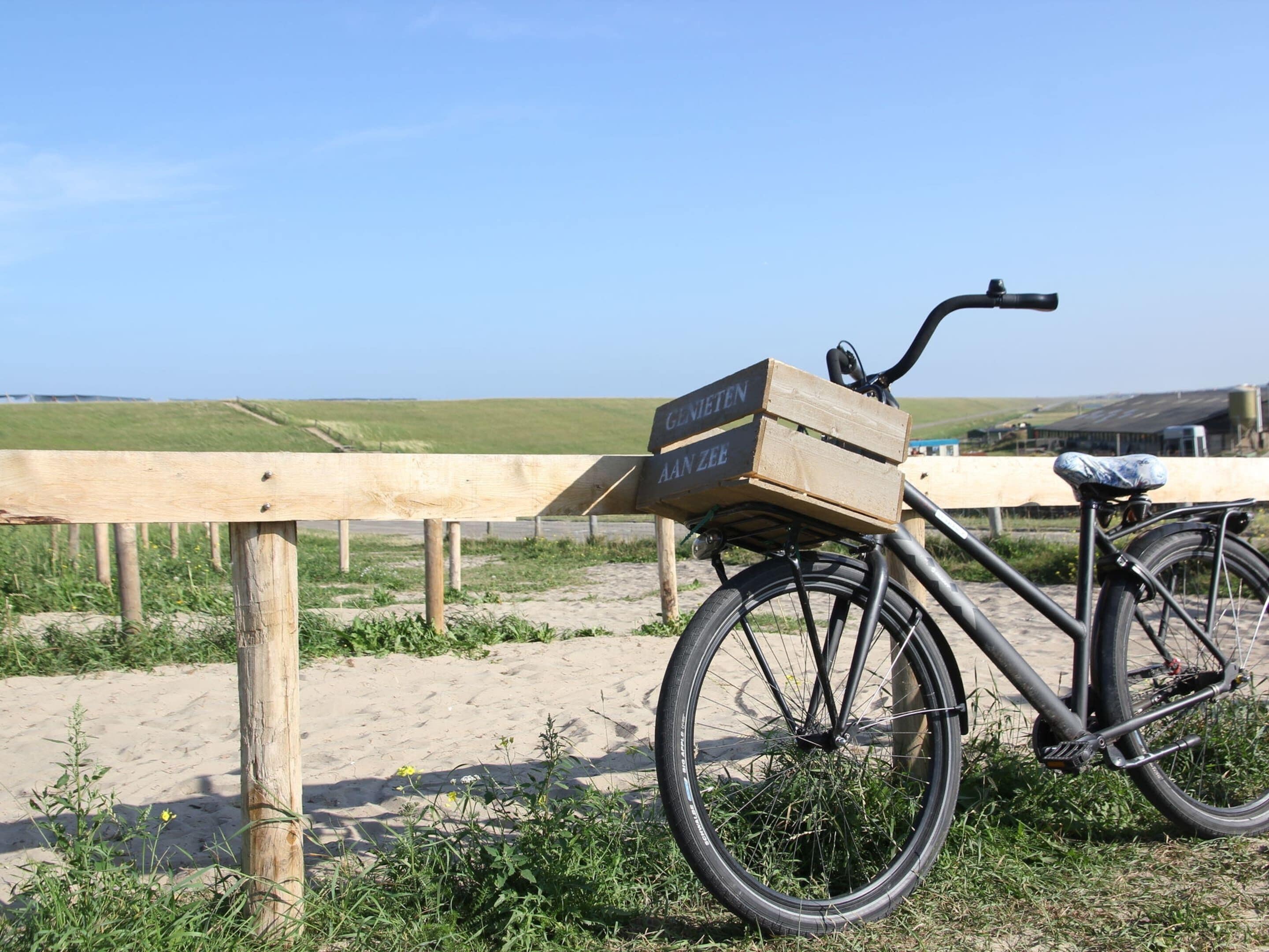 Un vélo avec une boîte en bois fixée à l’avant, garé sur une zone sablonneuse, avec une clôture en bois et de la verdure en arrière-plan.