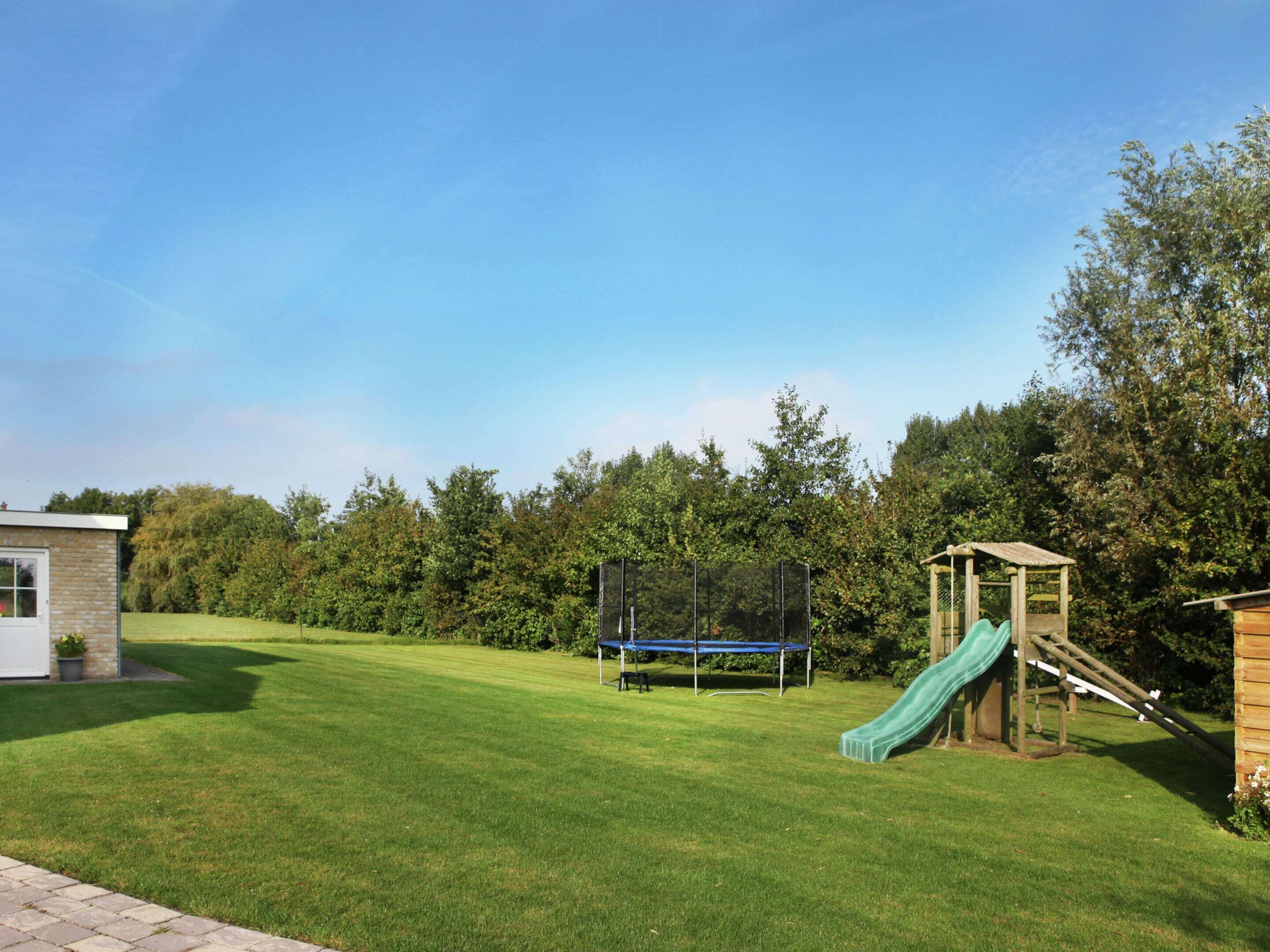 A grassy backyard with a trampoline, a small wooden playhouse with a slide, and a brick building.