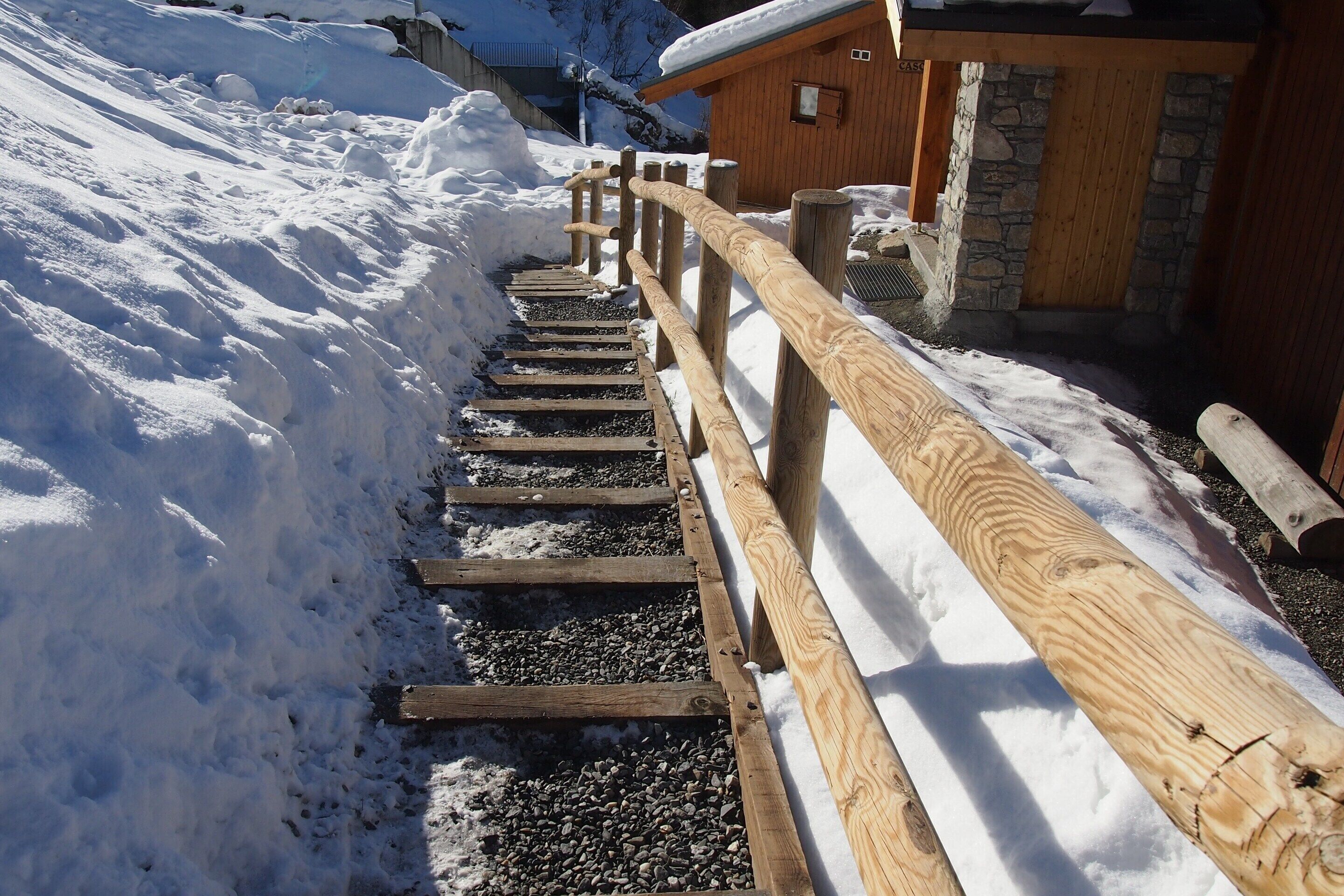 Un escalier en bois menant à un bâtiment à la façade en pierre, entouré de neige.