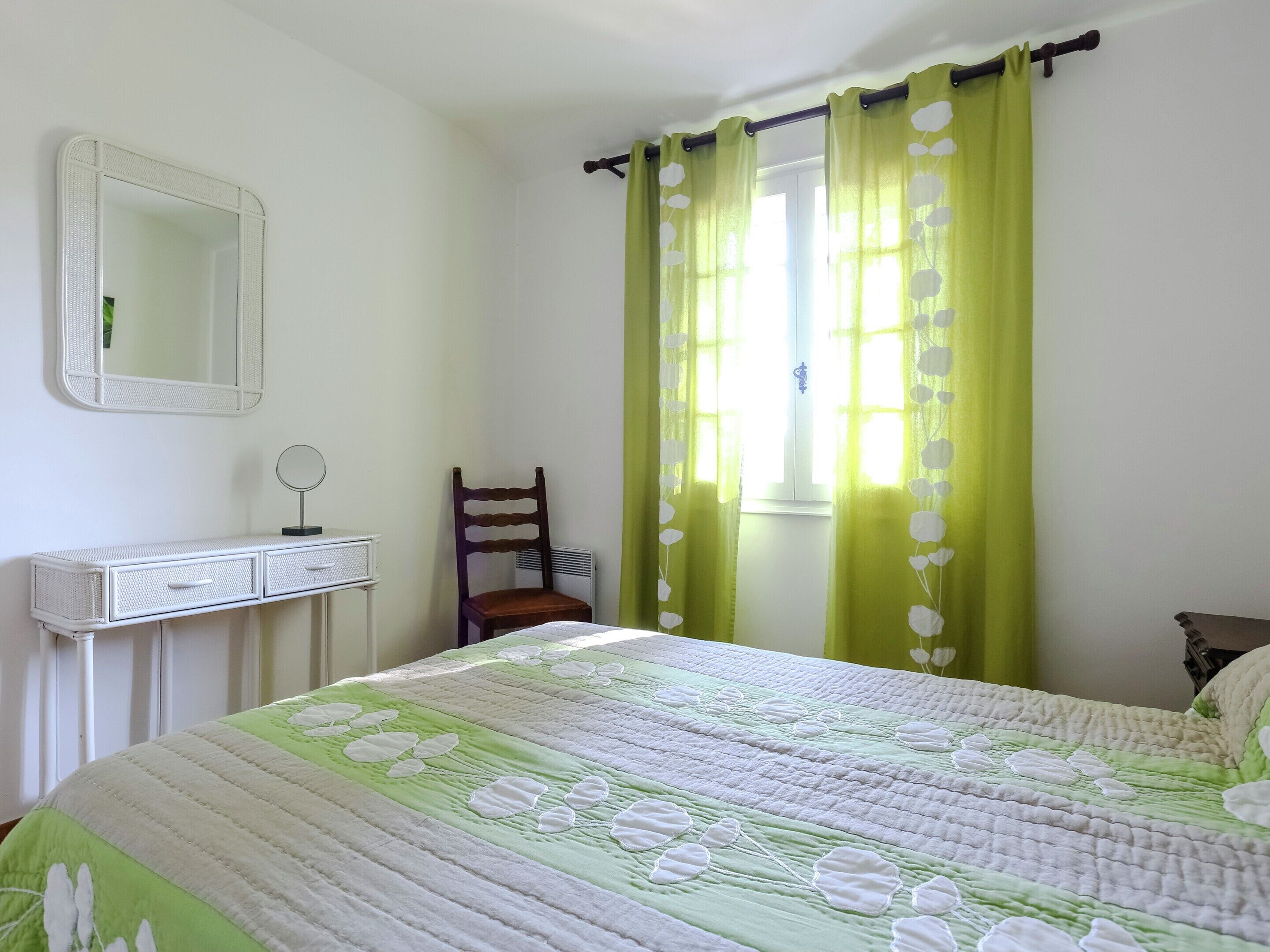 Une chambre à coucher avec un lit, une chaise, une commode, un miroir et une fenêtre avec des rideaux.