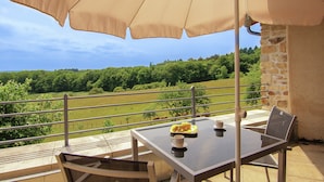 Ein Tisch mit Glasplatte, zwei Stühle, ein beiger Sonnenschirm und eine Ablage mit Essen und Tassen auf einem Balkon mit Blick auf eine grüne Landschaft.