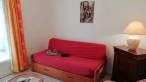 Een rode bank met pantoffels, een klein rond tafeltje met tijdschriften, een lamp op een houten bijzettafeltje, en een rieten stoel.