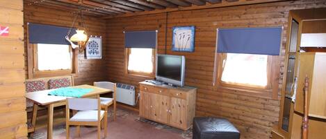 A cozy cabin interior with a wooden ceiling, a television, a dining area with a table and chairs, and a small kitchenette.