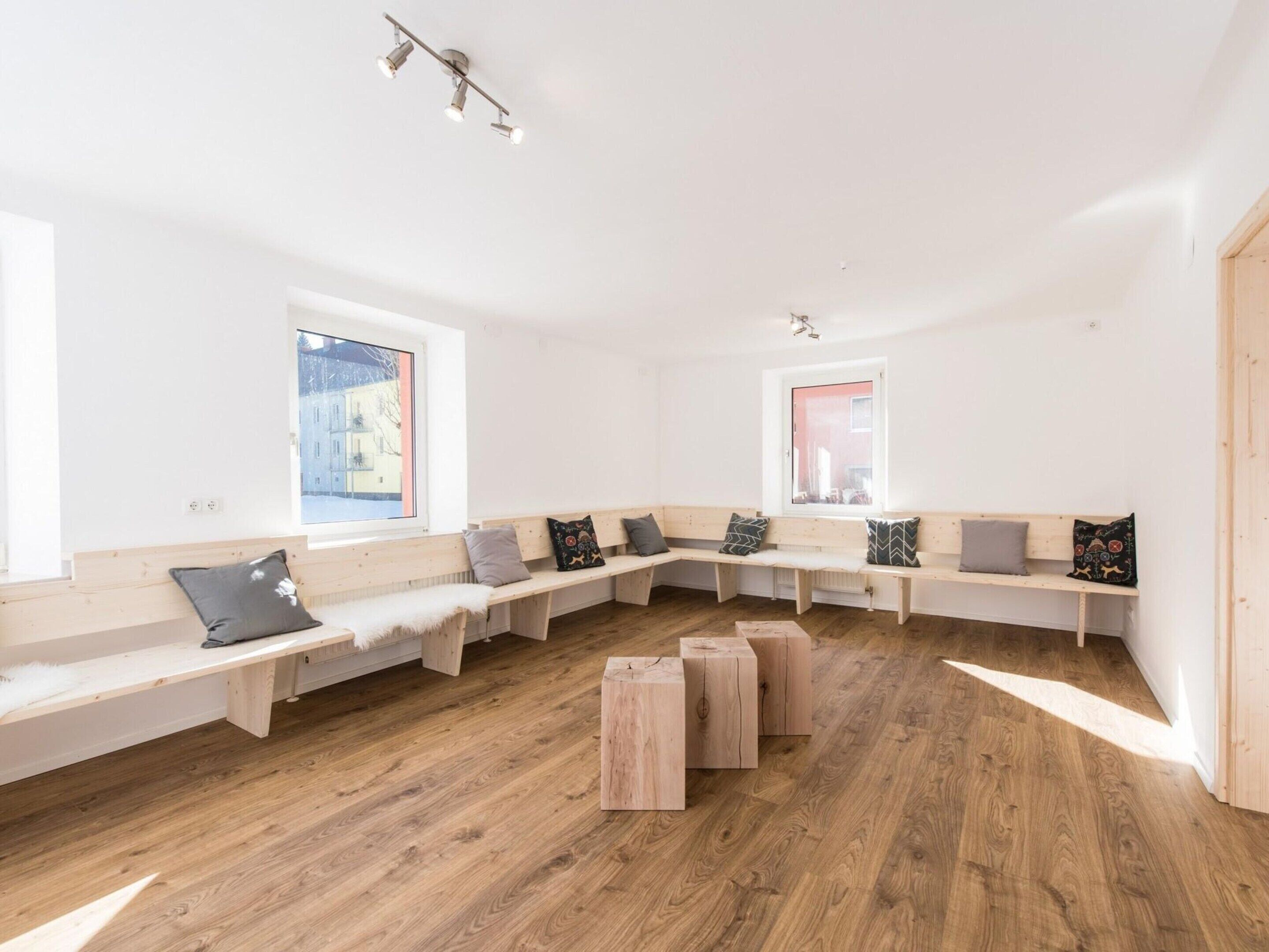 A modern, well-lit room with wooden benches and a central table, large windows with views of buildings, and a simple, clean design.
