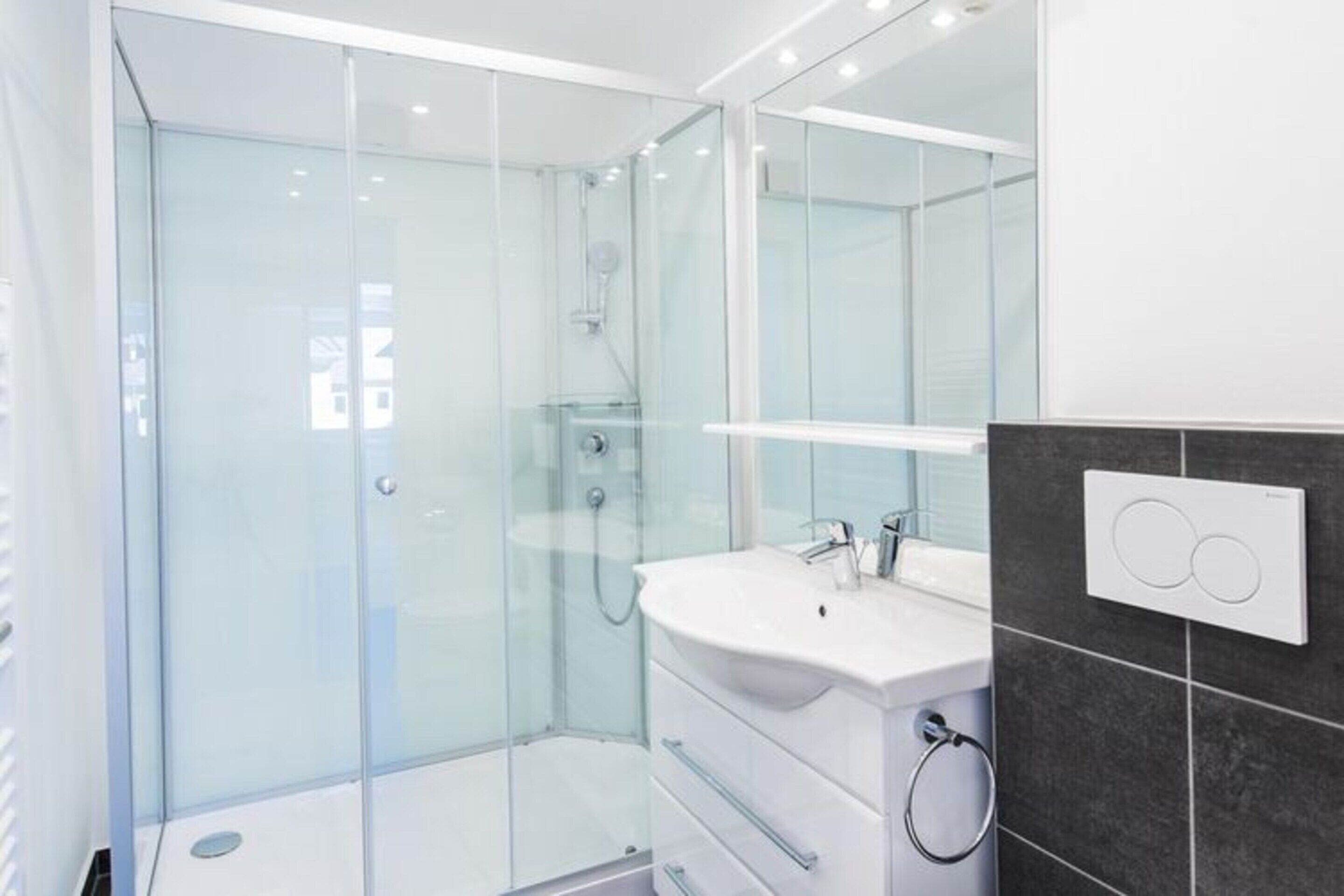 Une salle de bain moderne avec une douche à parois de verre, un lavabo blanc et un mur recouvert de carreaux foncés.