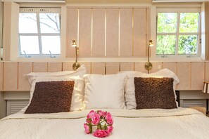 Una cama bien tendida con almohadas blancas y marrones, dos lámparas fijadas a la pared y un ramo de flores rosas sobre la cama.