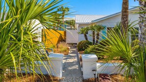 A pathway leading to a house with a white exterior and a yellow door, surrounded by palm trees and a clear blue sky. - 30ft from Perfect LAGUNA BEACH ~ Directly across the street from Beach Access #83 "Oleander West Cottage" (Panama City Beach)