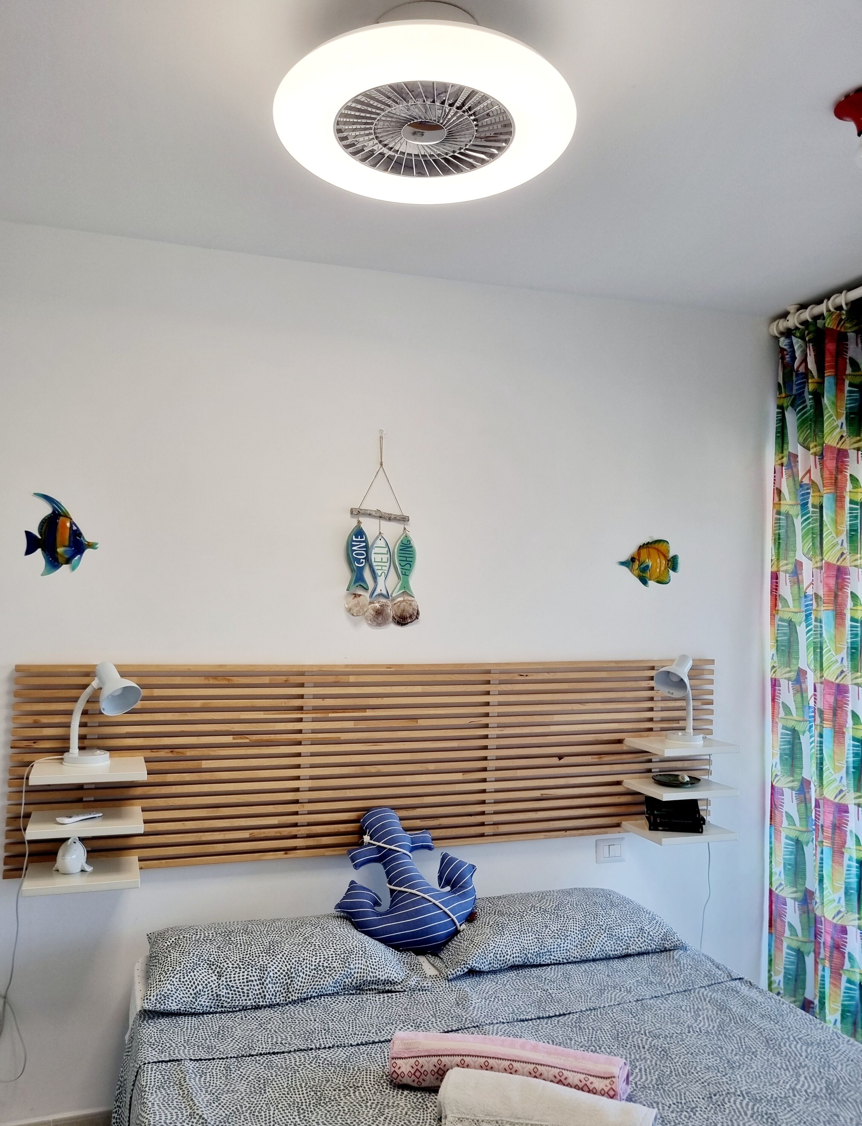 A bedroom with a wooden slat headboard, a bed with patterned bedding, a hanging fish decoration, and a colorful curtain.