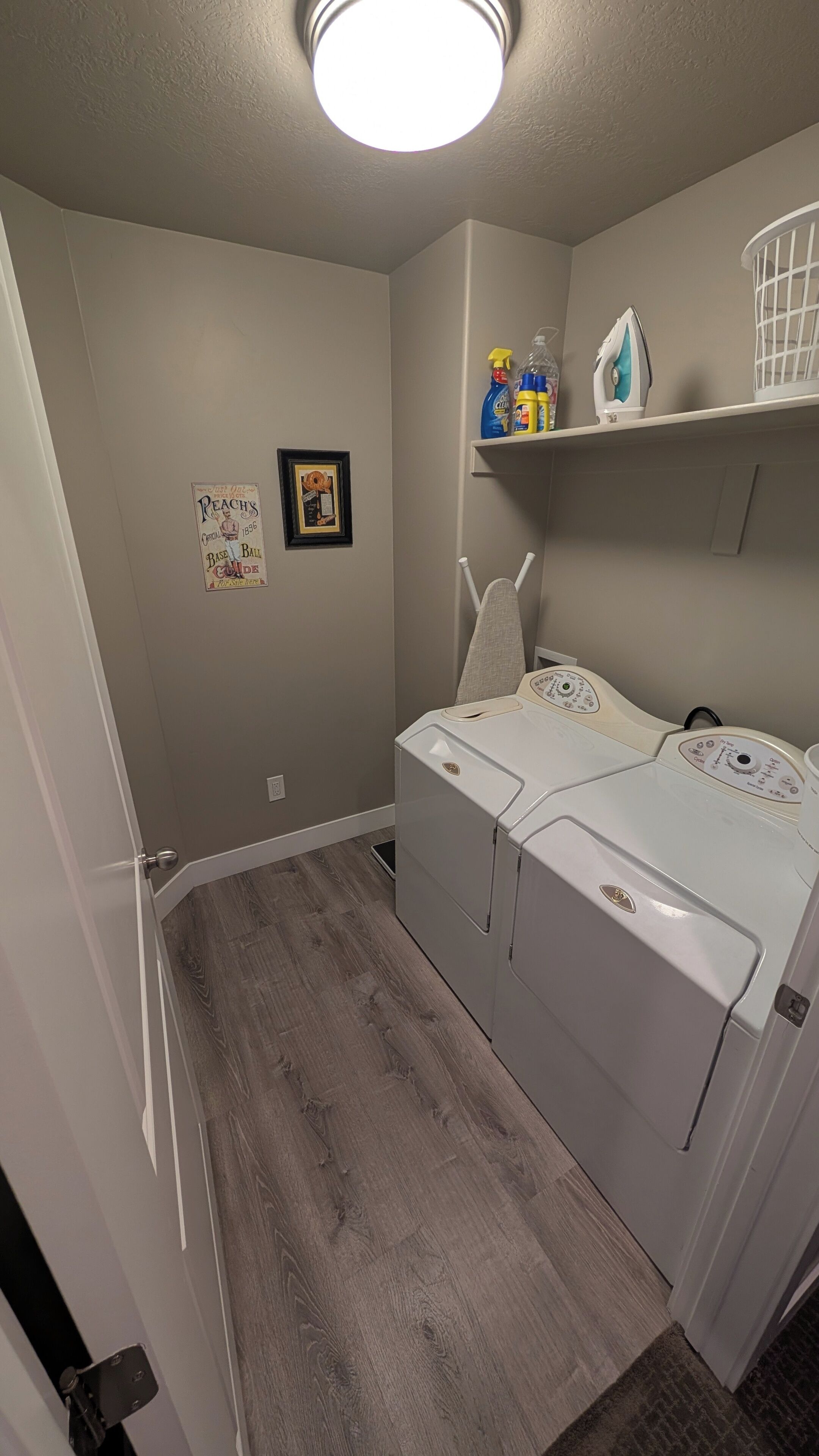 A laundry room with a dryer, ironing board, and shelves with cleaning supplies.