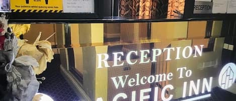 A reception area with a 'Welcome To Pacific Inn Hotel' sign, a 'Keep Your Distance' sign, and a reception desk.