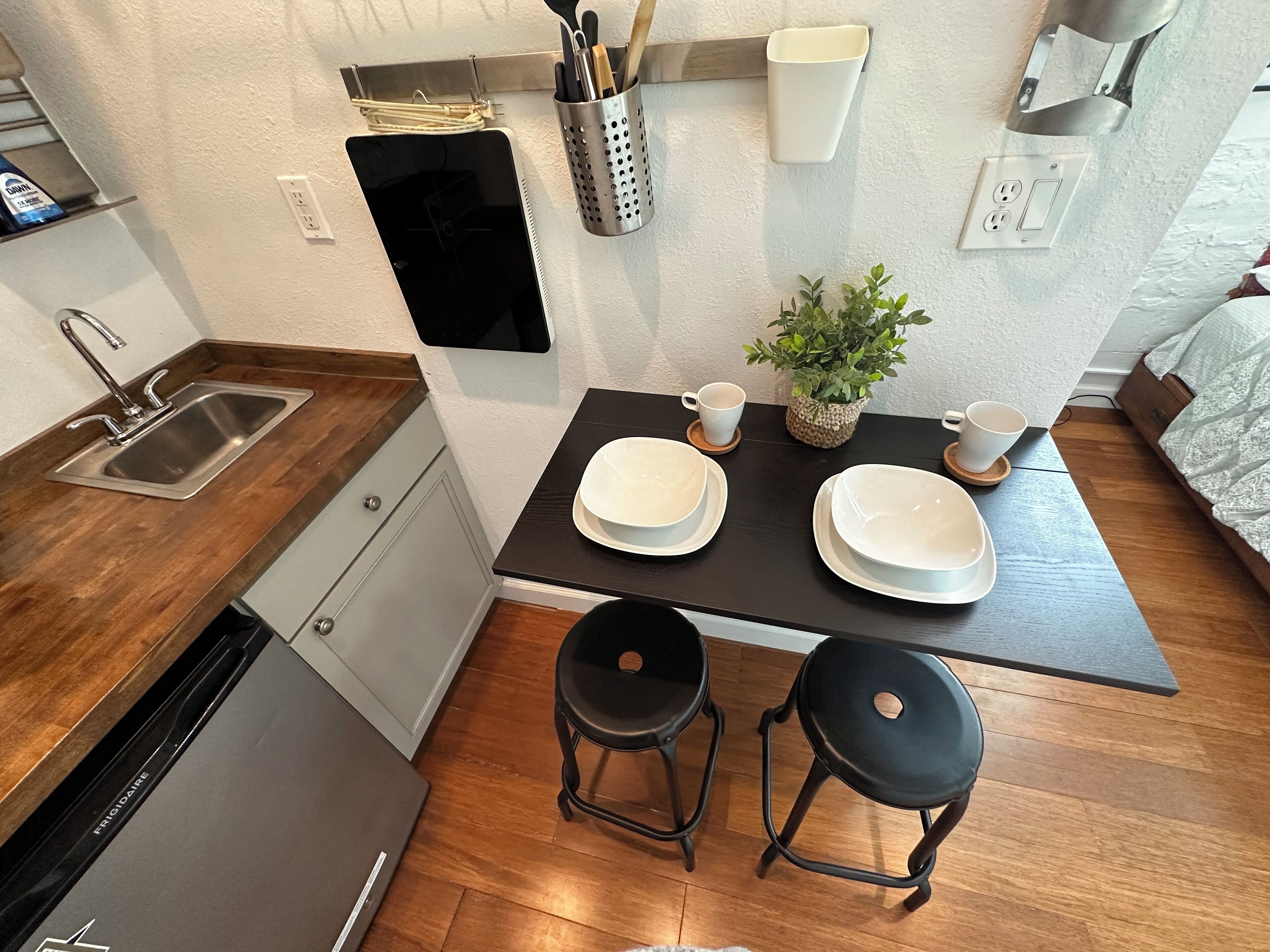 A compact kitchen with a sink, a small refrigerator, and a dining area with two stools and a table set with plates and a cup.
