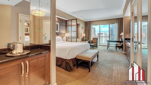 Ein Hotelzimmer mit einem großen Bett, einem Schreibtisch, einem Sessel und einem Esstisch, mit Blick auf die Stadt.