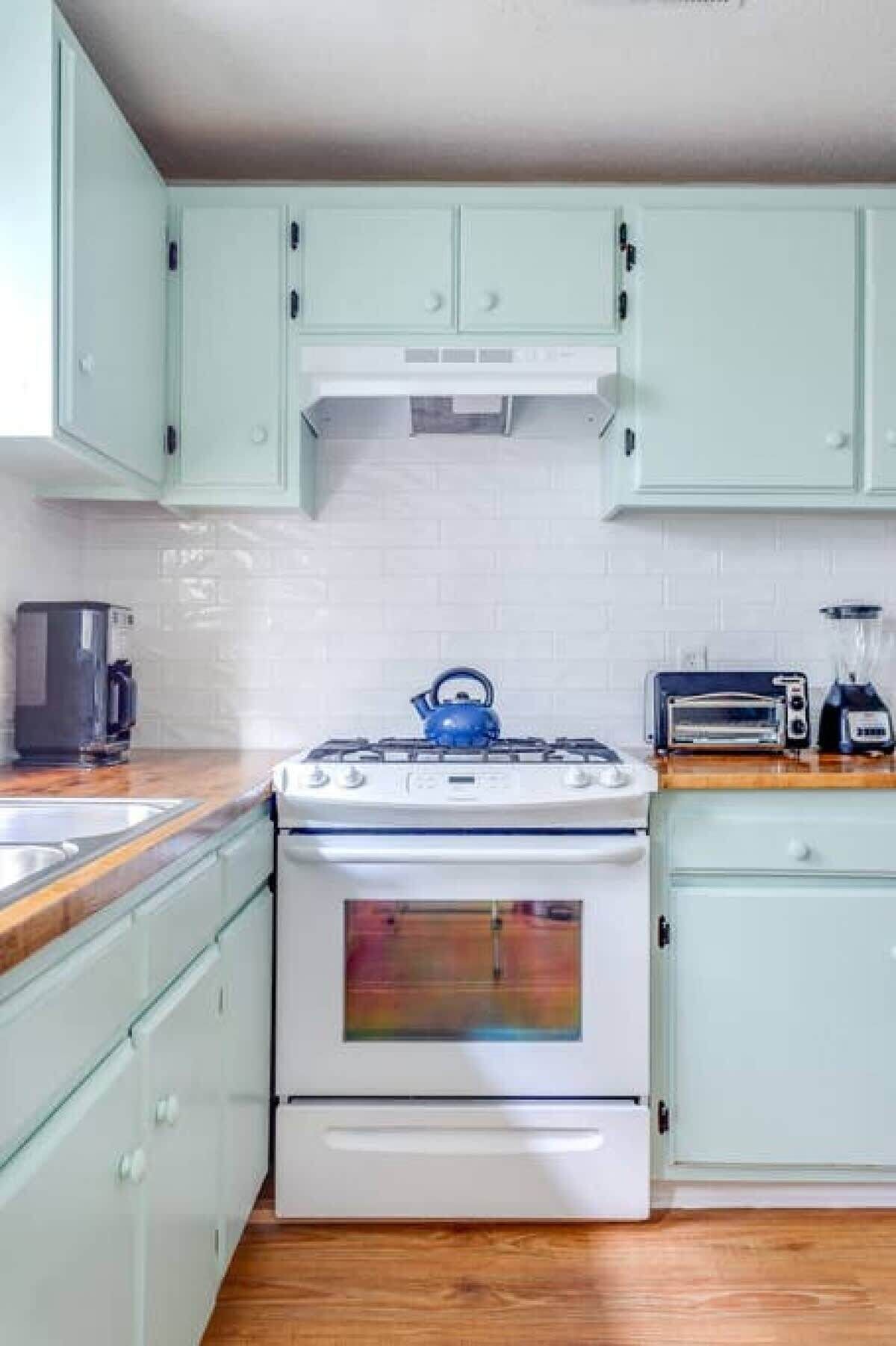 A kitchen with light green cabinets, a white stove, a blue kettle, a coffee maker, a toaster, and a blender.