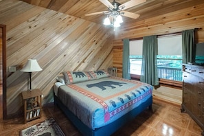 A bedroom with a sloped ceiling, a bed with a bear-themed bedspread, a wooden dresser, a nightstand with a lamp, and a window with curtains. - Franklin, NC Cabin; Hot Tub; Mountain View; Starlink (Franklin)