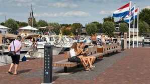 Un port de plaisance avec des bateaux amarrés, le clocher d’une église en arrière-plan, et des personnes assises sur des bancs.