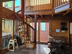 Interior de una cabaña de madera con una escalera espiral, una mesa de comedor y una zona de cocina.