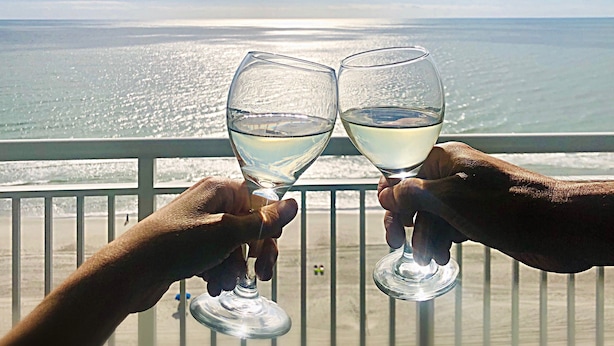 Deux mains tenant des verres de vin, avec l’océan en toile de fond.