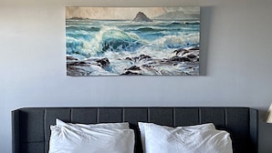 Une chambre à coucher avec un lit, deux oreillers blancs, une tête de lit sombre et un tableau représentant un paysage marin accroché au mur.