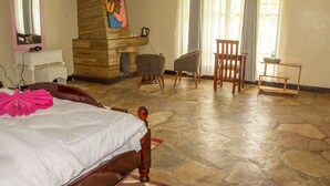 A bedroom with a stone floor, a bed with a wooden headboard, a chair, a small table, and a painting on the wall. - Ingagi Lodge, UNIT 1, Single room (Kinigi,)