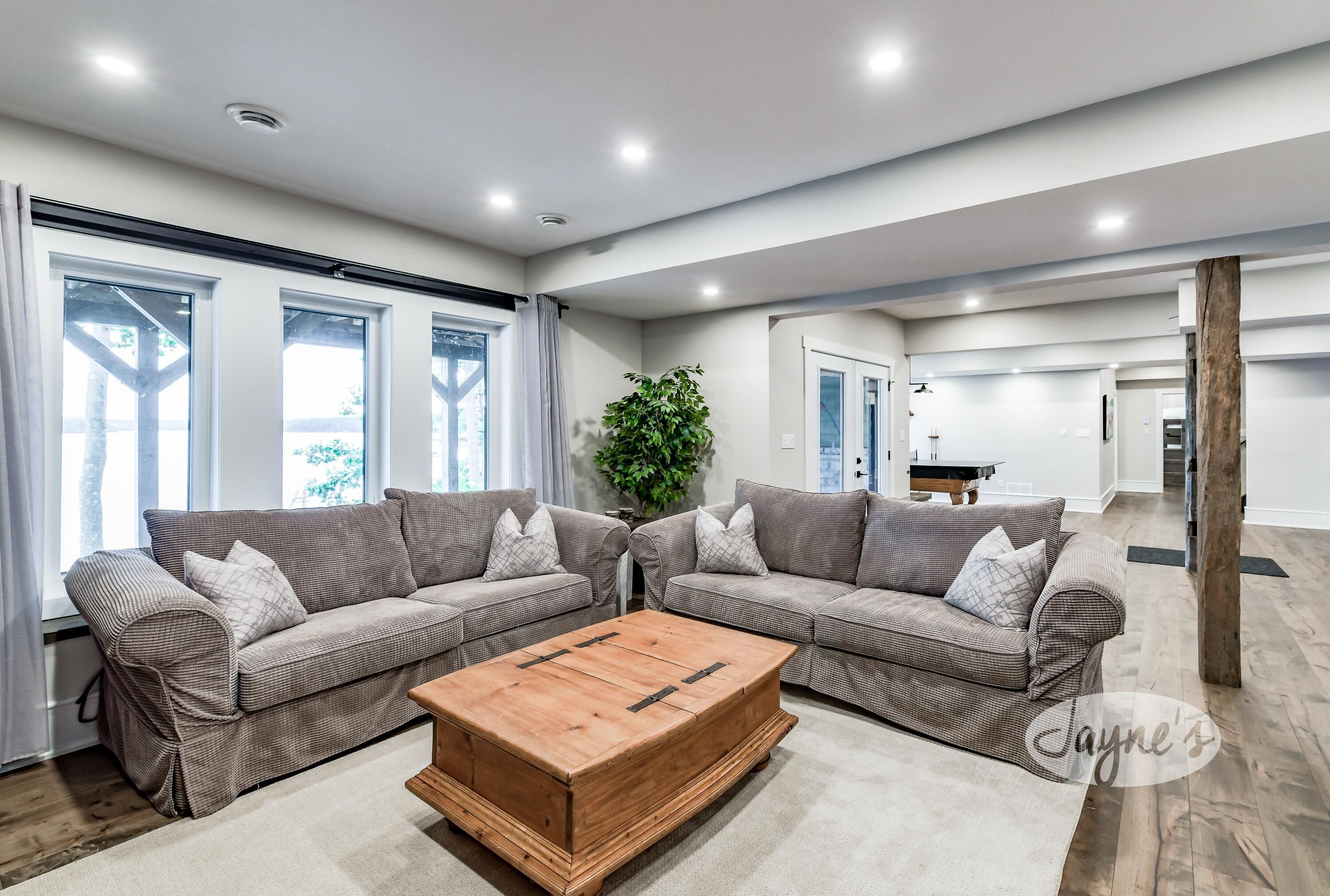 A modern living room with two grey sofas, a wooden coffee table, large windows with curtains, and a plant.