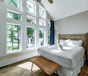 A bedroom with a large bed, multiple windows, a wooden bench, and a nightstand with a lamp.
