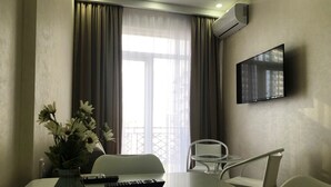 A hotel room with a dining table, two chairs, a vase with flowers, a flat-screen TV mounted on the wall, and a large window with curtains. - D&D Lux Apartment 96. Clean, bright and modern.  (Batumi)