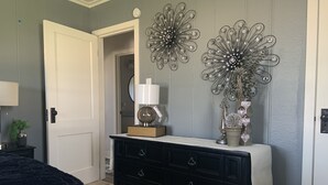 A bedroom with a black dresser, a white lamp, decorative wall art, and a bed with a white cover.