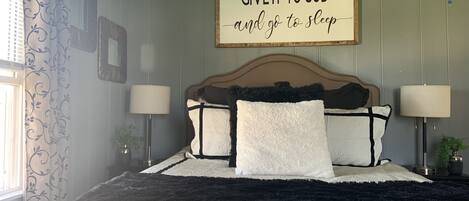 A bedroom with a bed, two bedside lamps, a wall sign, and a window with curtains.
