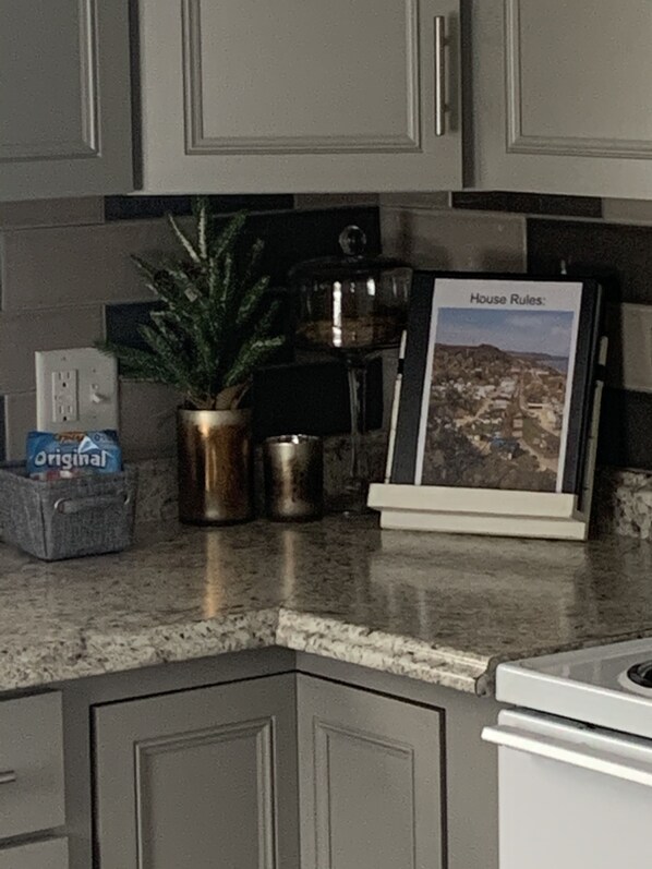 A kitchen with granite countertops, a potted plant, a coffee maker, and a framed picture titled "House Rules".