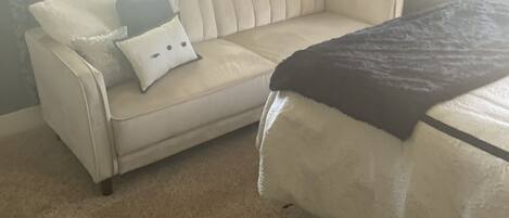 Une pièce avec un canapé beige, un lit recouvert d’une couverture foncée et une fenêtre avec des rideaux.