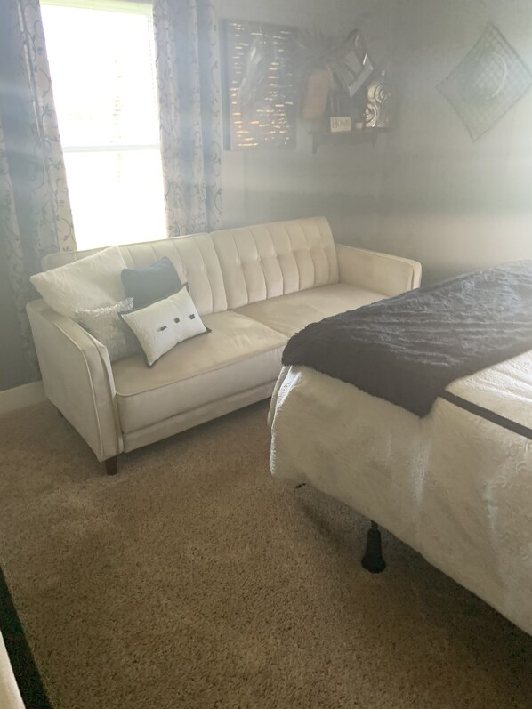 A room with a beige sofa, a bed with a dark blanket, and a window with curtains.