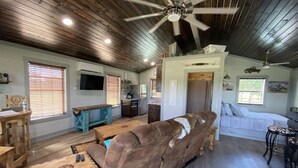 A spacious living area with a brown leather sofa, wooden furniture, a ceiling fan, and a kitchen area with a refrigerator and stove.