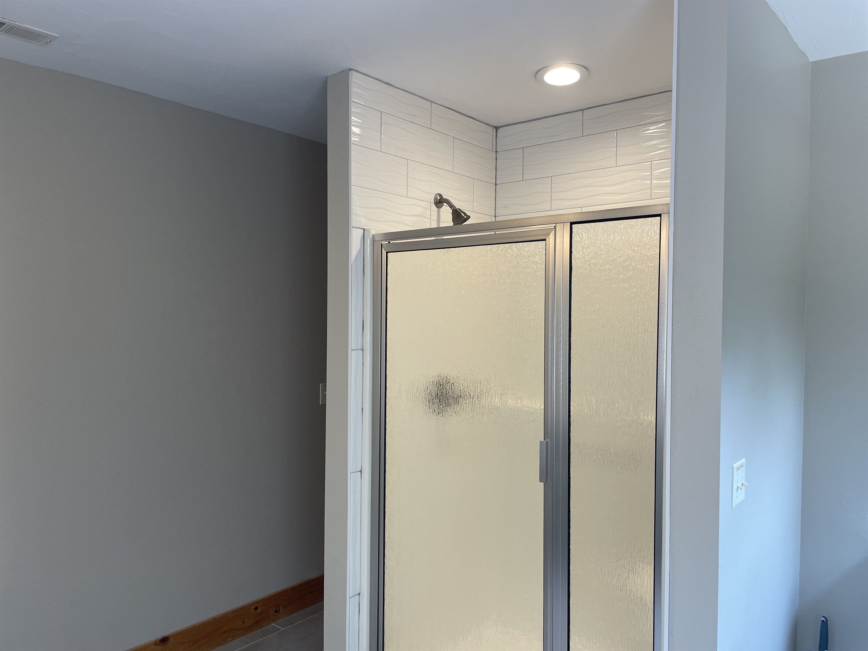 A bathroom with a glass shower door, white tiled walls, and a wooden baseboard.