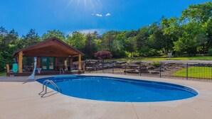 A wooden cabin with a pool, surrounded by a fenced area and trees.