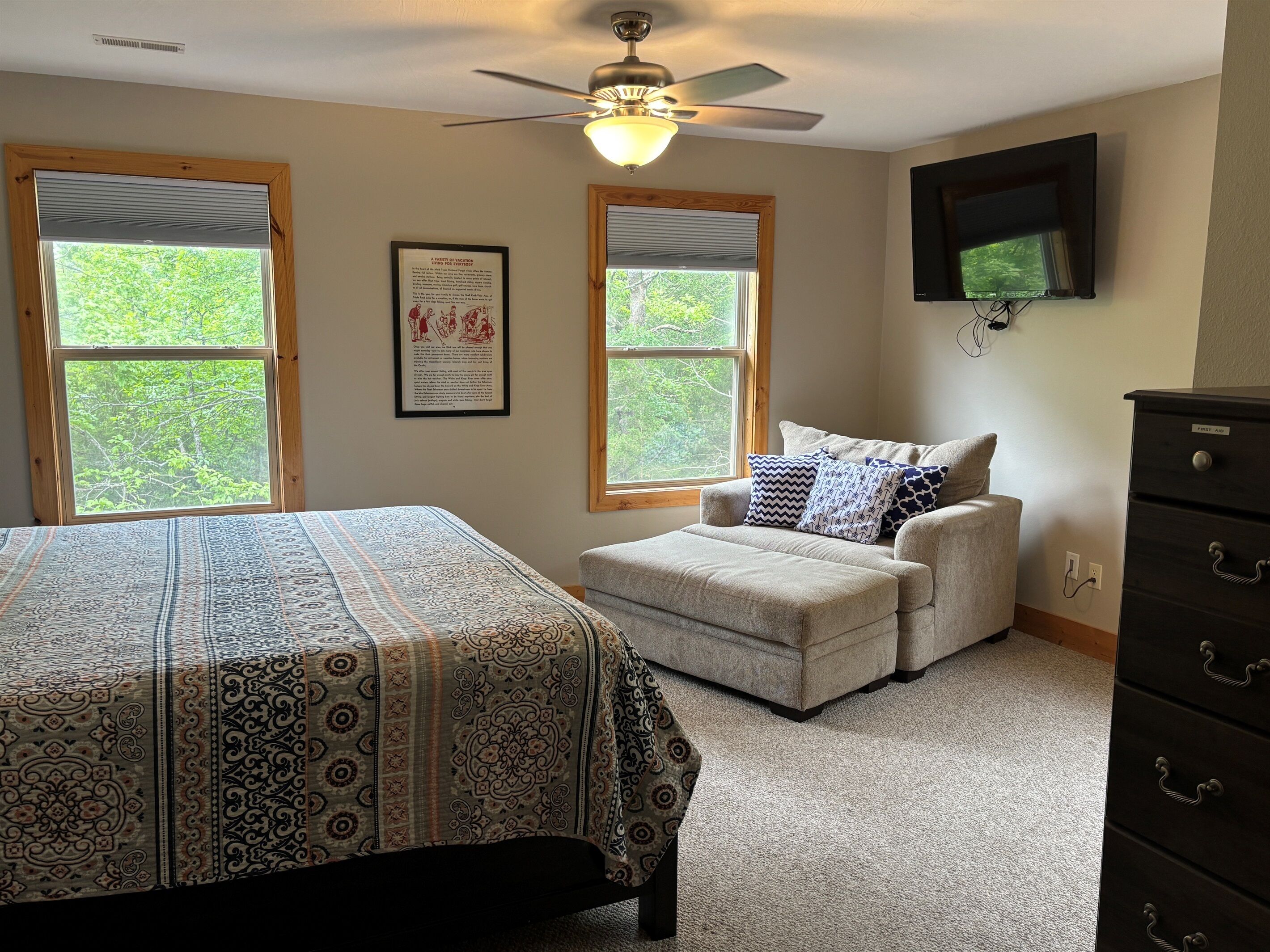 A bedroom with a bed, an ottoman, a ceiling fan, two windows, and a dresser.