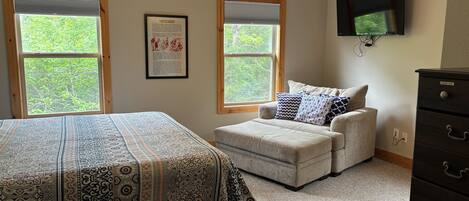 A bedroom with a bed, an ottoman, a ceiling fan, two windows, and a dresser.