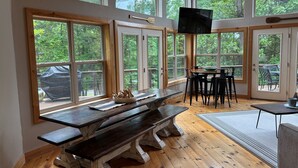 A spacious room with large windows, a wooden bench, a dining table, and a TV mounted on the wall.