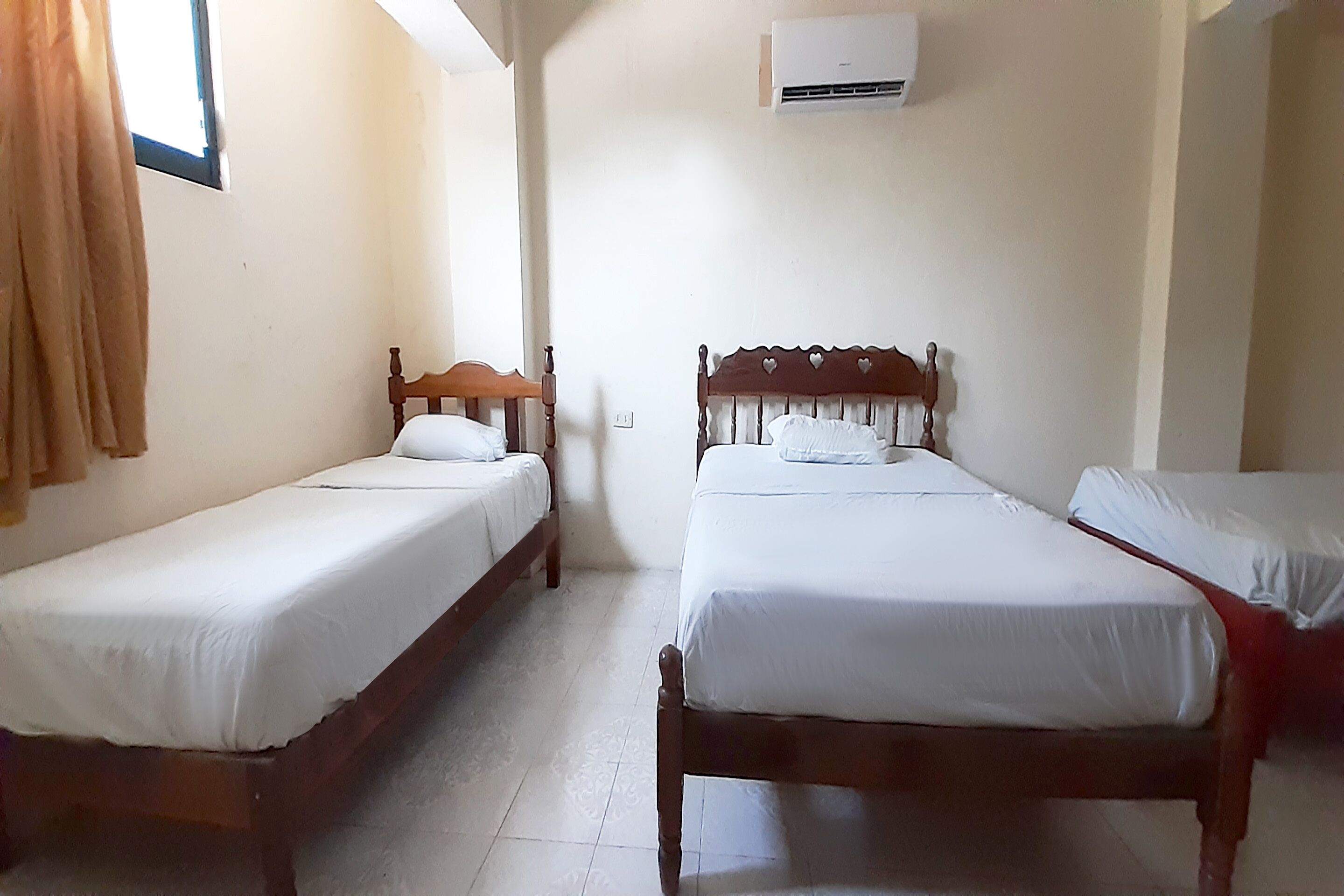 A room with two wooden beds, white bedding, a window with curtains, and an air conditioning unit on the wall.