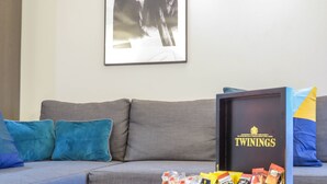 A living room with a grey sofa, blue cushions, a framed picture on the wall, and a box of Twinings tea bags on a table. - Tudors eSuites City Centre Houses with Garden (Birmingham)