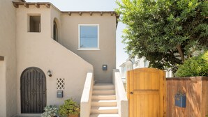 Une maison aux murs recouverts d’un enduit beige, avec une porte en bois, un escalier en béton et un pot de fleurs.