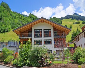 A two-story building with a wooden facade and multiple balconies, surrounded by lush greenery and a mountainous backdrop. - Alpina Appartements (Schroecken)