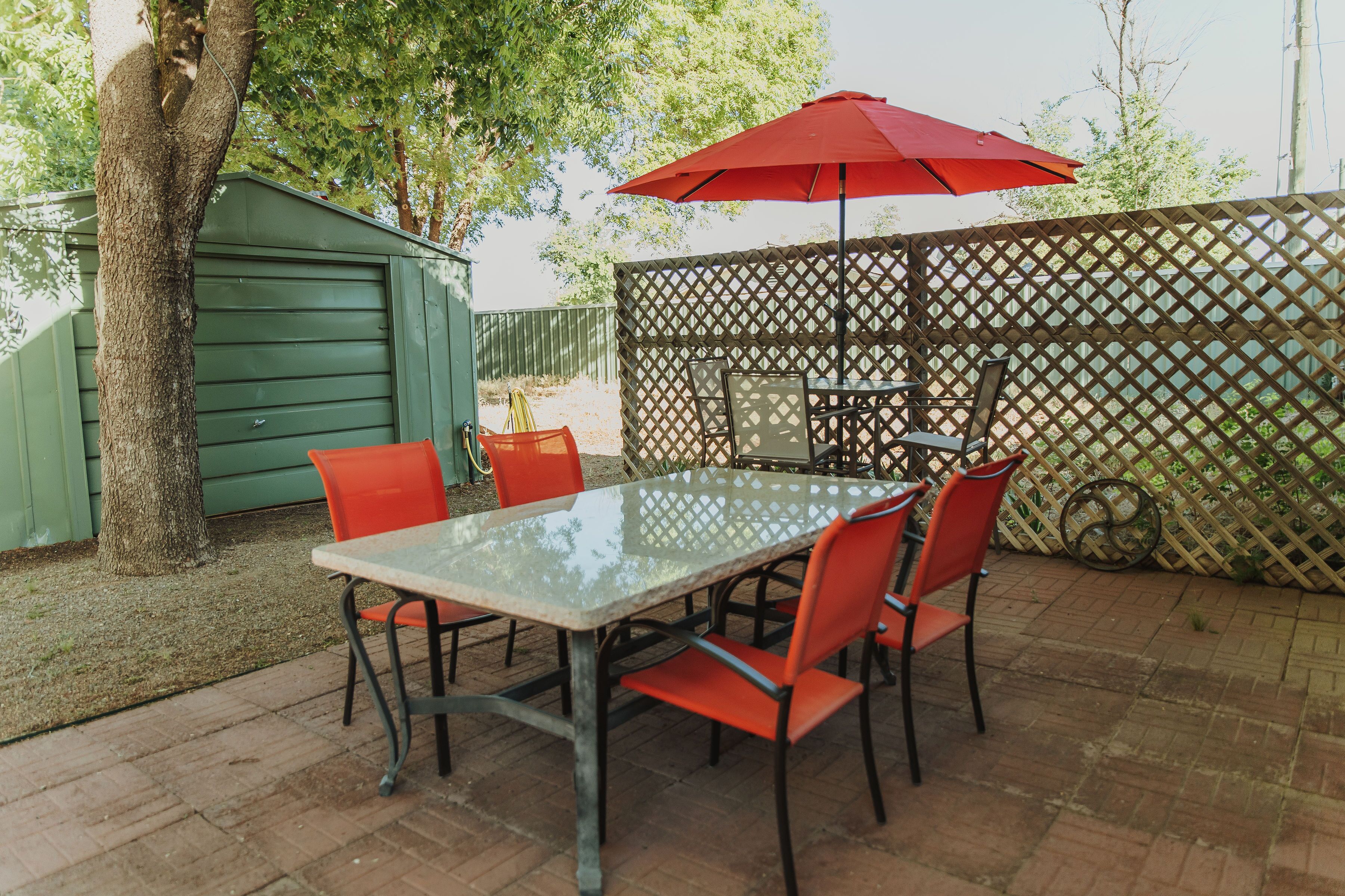An outdoor patio with a glass table and red chairs, a red umbrella, and a wooden fence.