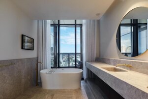 A modern bathroom with a freestanding bathtub, a large mirror, and a city view through floor-to-ceiling windows.